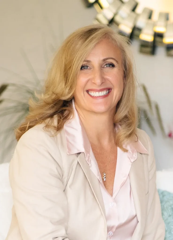 1a Smiling woman with blonde hair in a light beige blazer and pink blouse, indoors with plants in the background.